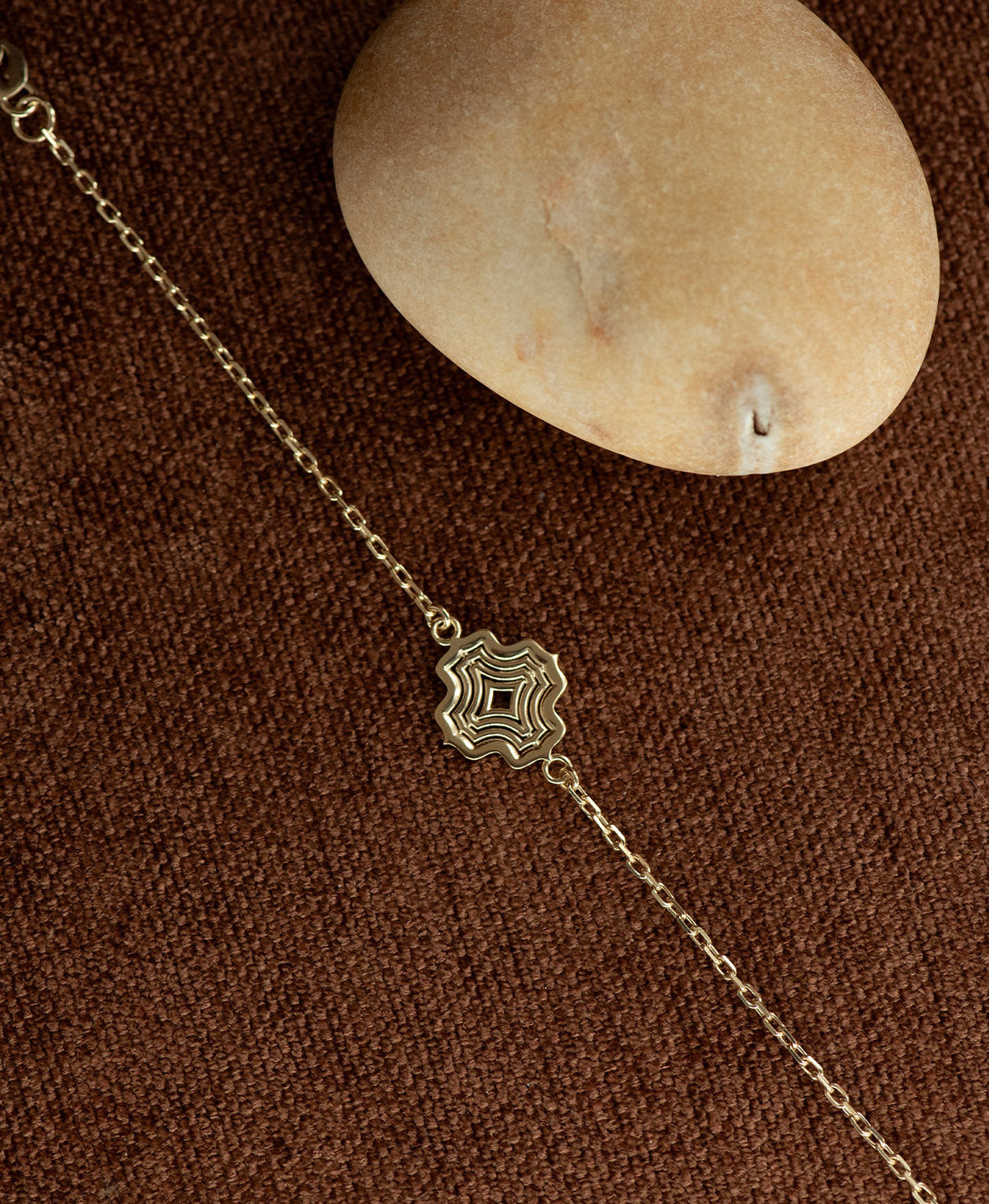 The Alma Bracelet featuring a gold pendant with a concentric wave design, resting next to a cream stone on a textured brown fabric background.