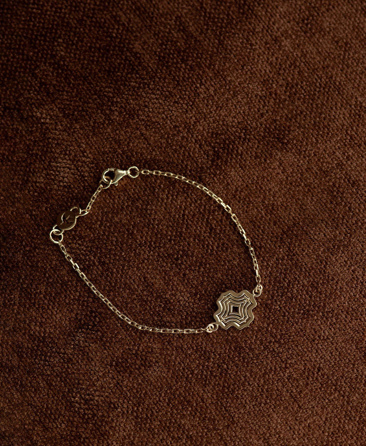 The Alma Bracelet featuring a gold pendant with a concentric wave design, on a textured brown fabric background.