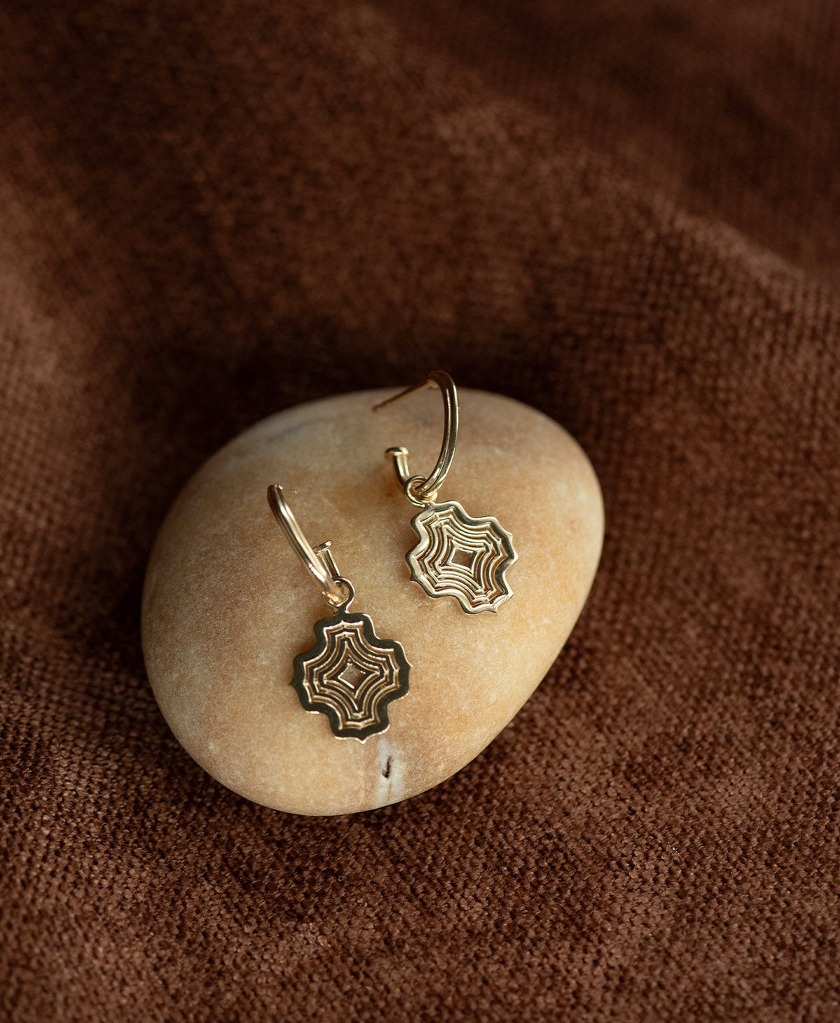 The Alma Hoops, featuring a gold pendant with a concentric wave design suspended from a hoop, displayed on a smooth beige stone against a textured brown fabric background.