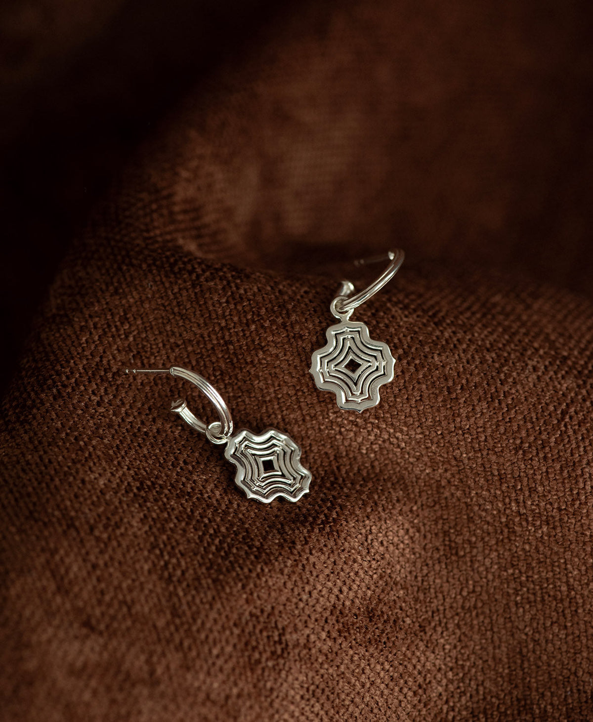 The Alma Hoops, featuring a silver pendant with a concentric wave design suspended from a hoop, displayed against a textured brown fabric background.