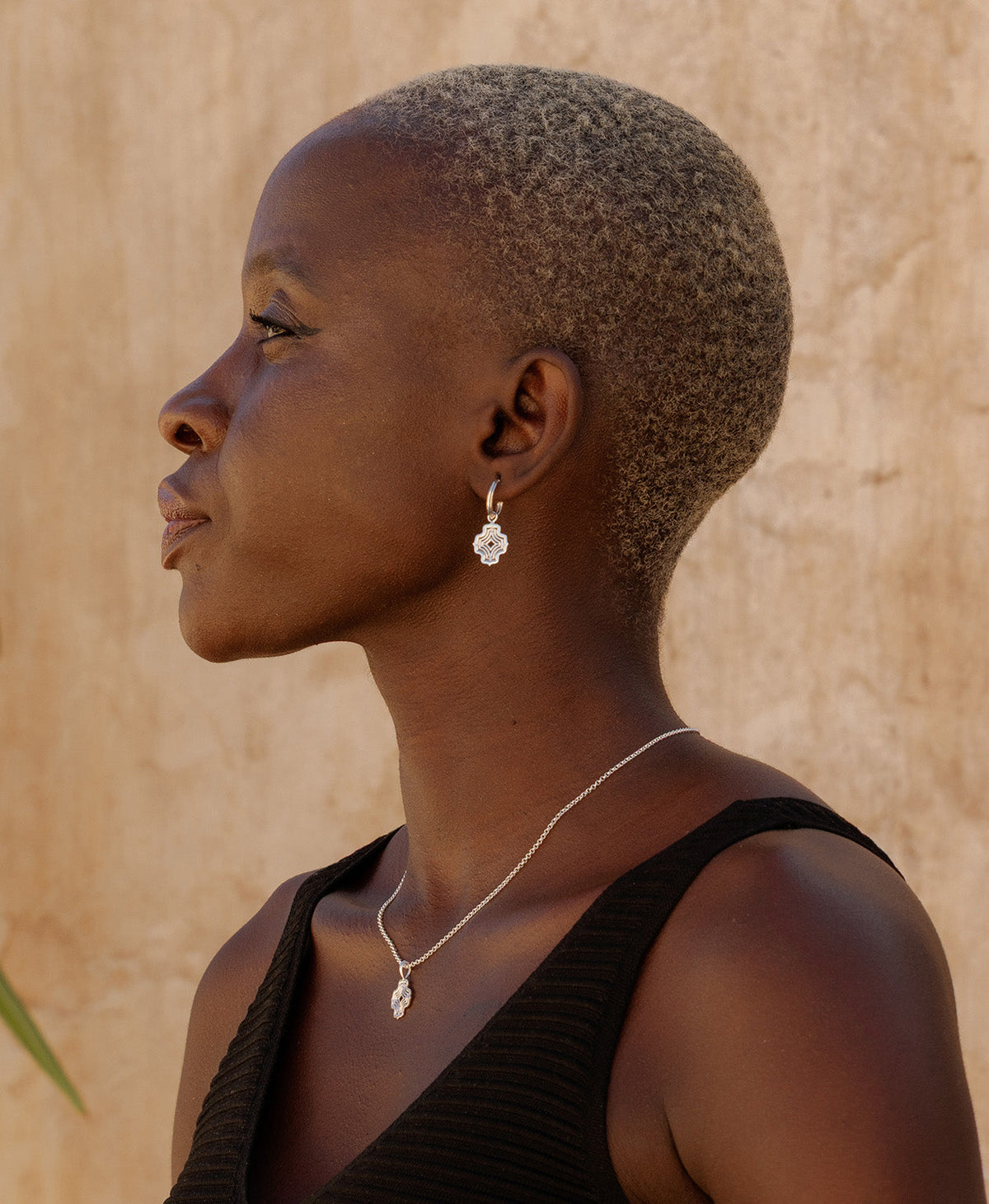 A close-up side profile of a person wearing the Alma Hoops, featuring a gold pendant with a concentric wave design, suspended from a hoop, paired with a silver pendant necklace.