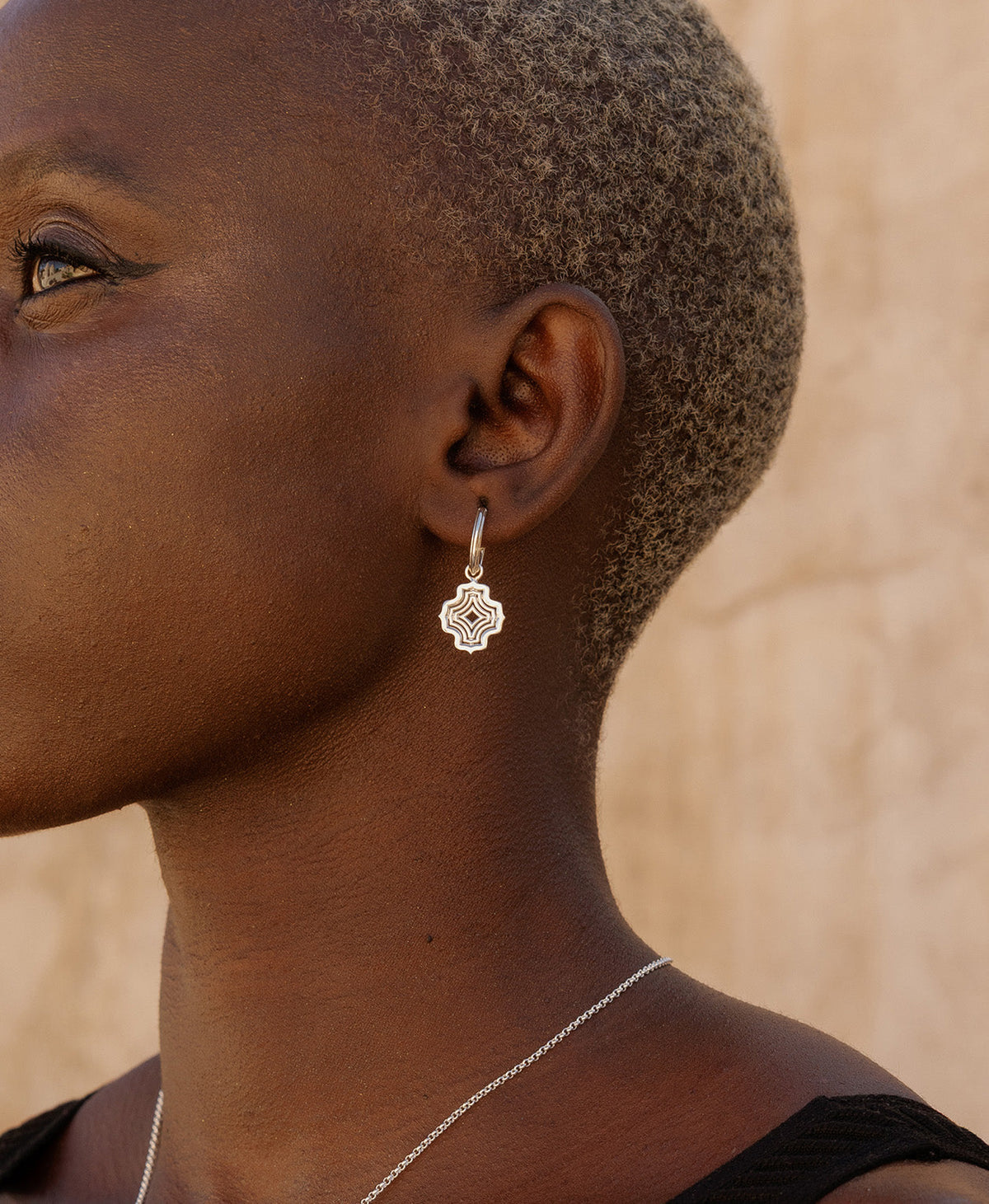 A close-up side profile of a person wearing the Alma Hoops, featuring a silver pendant with a concentric wave design, suspended from a hoop.