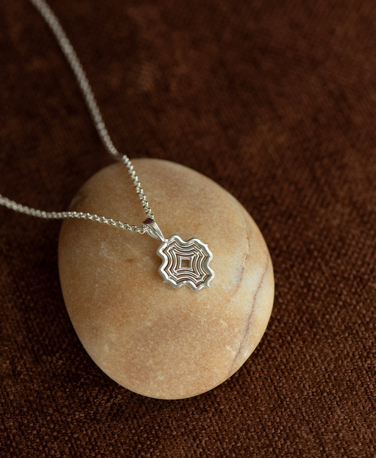 The Alma Necklace featuring a silver pendant with a concentric wave design, layered over a cream stone on a textured brown fabric background.