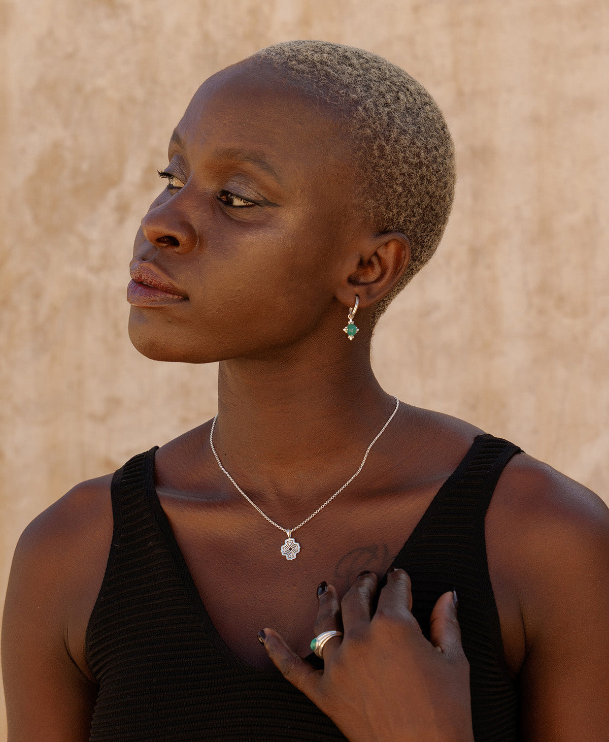A person wearing the Nora Necklace, a silver pendant with a concentric wave design, suspended from a fine belcher chain, paired with silver emerald earrings.