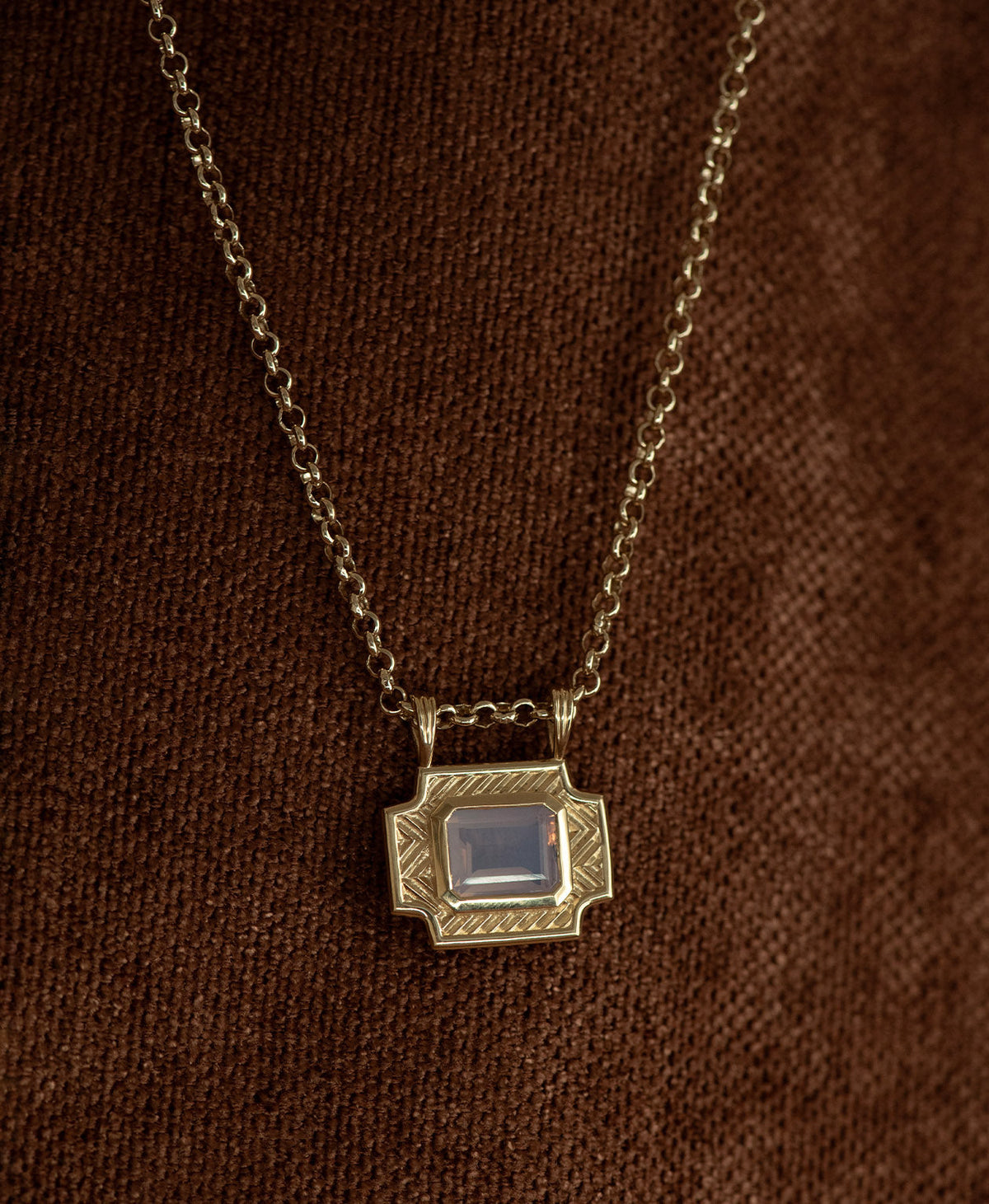 A gold pendant with a rectangular lavender quartz stone, handing on a belcher chain against a textured brown background.