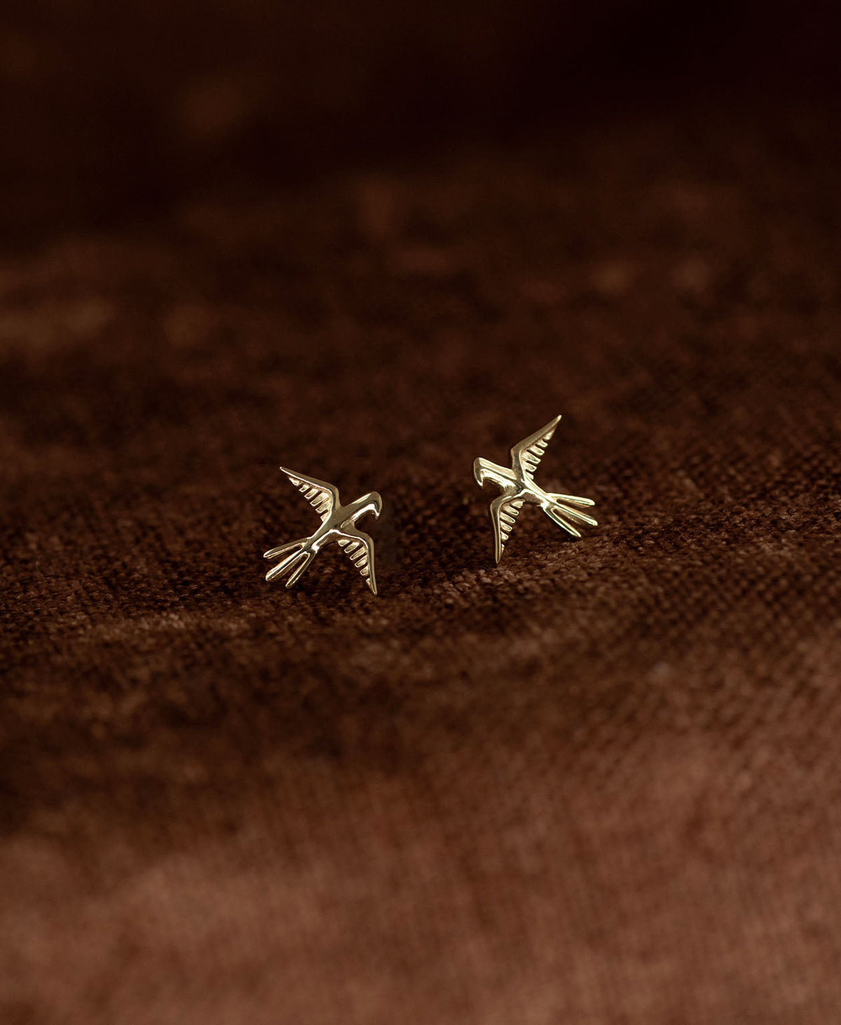 A pair of gold bird stud earrings with detailed feather and wing textures, displayed on a dark brown textured fabric background.