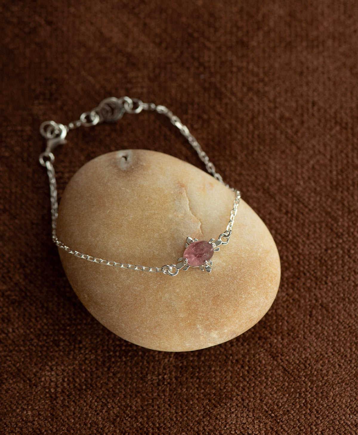 The silver Farah Bracelet featuring a cabochon cut pink tourmaline, layered over a cream stone on a textured brown fabric background.