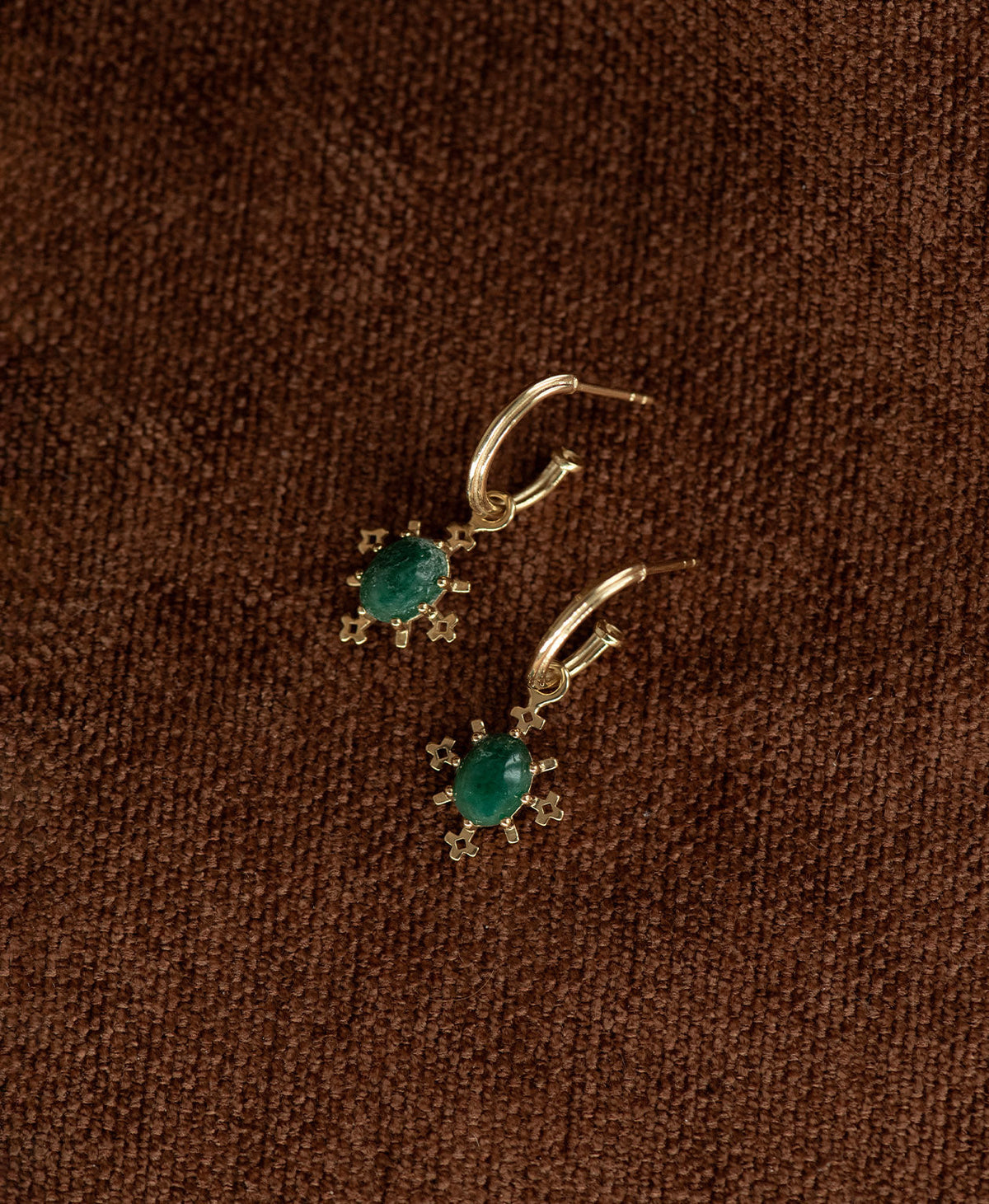 A pair of Farah gold drop earrings with emerald gemstones, displayed against a dark brown textured background.