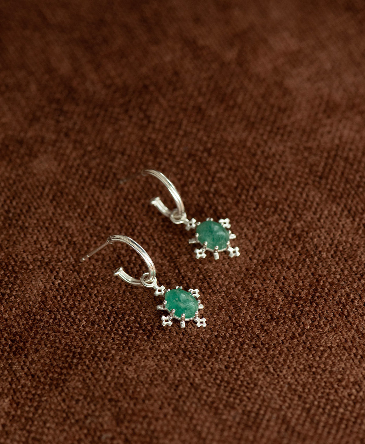 A pair of Farah silver drop earrings with emerald gemstones, displayed on a textured brown fabric background.
