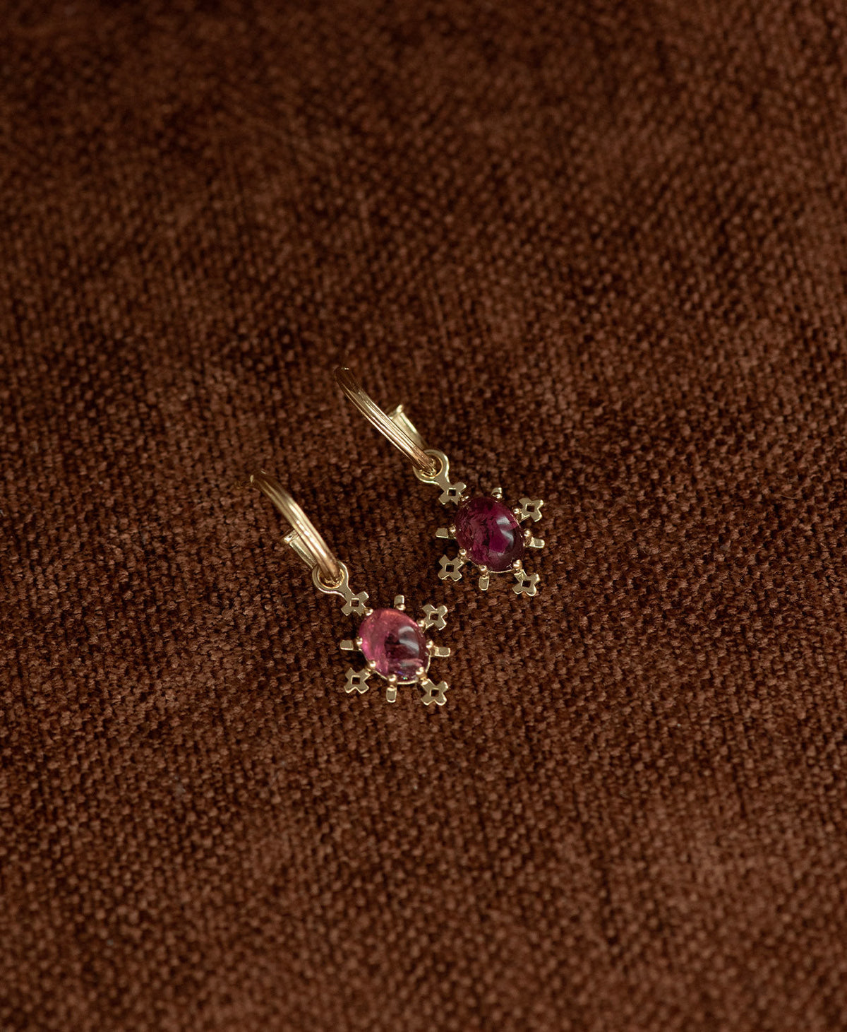 A pair of Farah gold drop earrings with pink tourmaline gemstones, displayed on a textured brown fabric background.