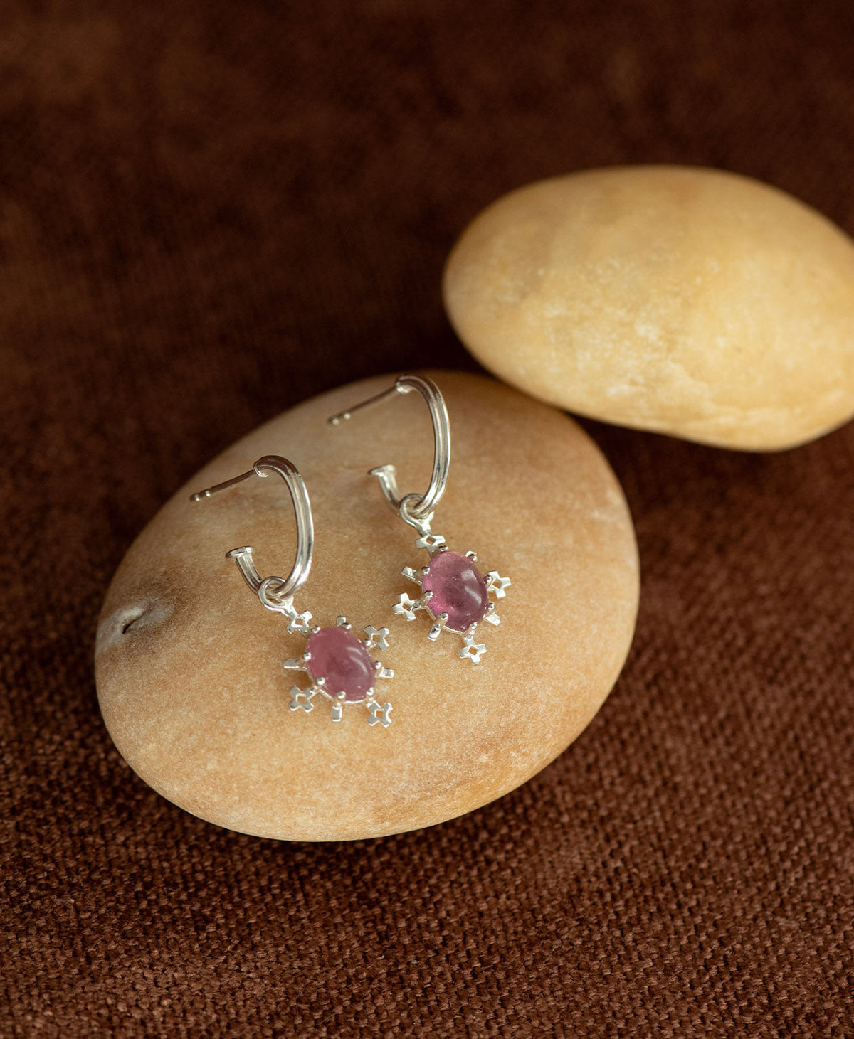 A pair of Farah silver drop earrings with pink tourmaline gemstones, displayed on a stacked smooth cream coloured stone against a dark brown textured background.