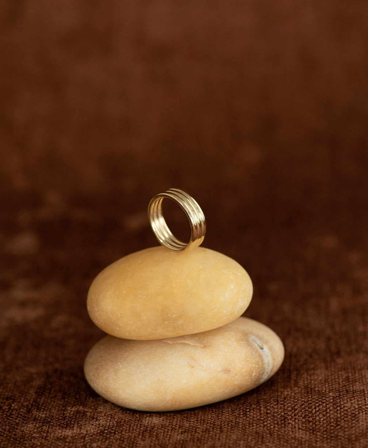 The gold Irma Stacker Ring, formed from three connected bands, layered closely, rests on stacked cream stones on a textured brown background.