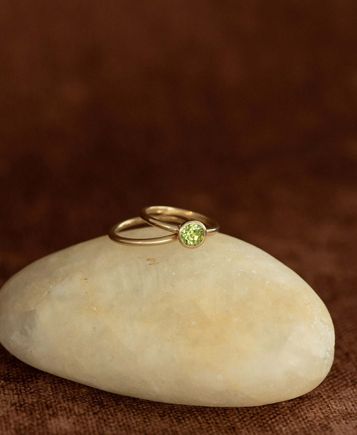 The smooth, curved Selma stacking ring in 22k gold vermeil, rests on a cream stone on a textured brown background, layered with a gold solitaire peridot ring.
