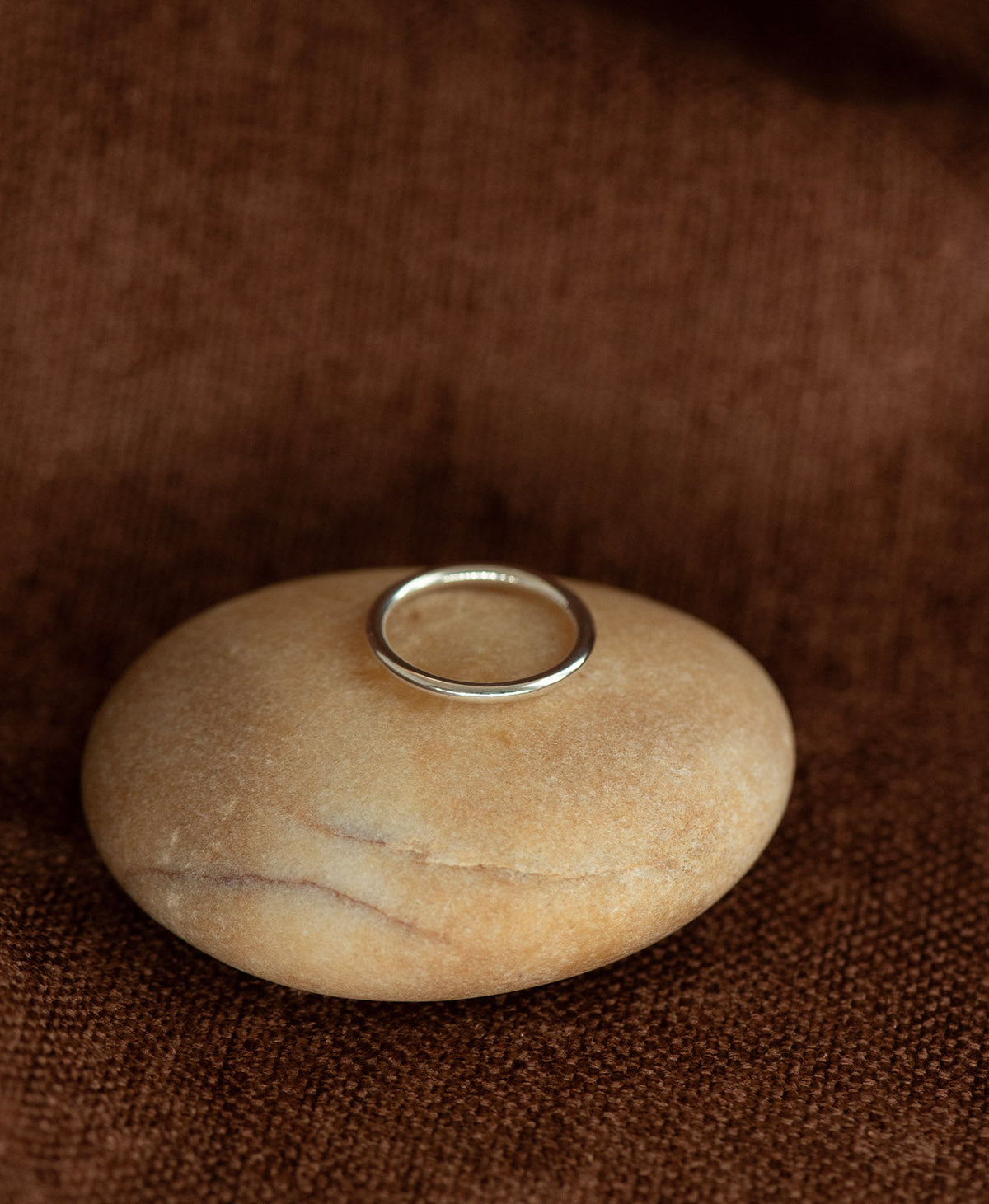 The smooth, curved Selma stacking ring in 925 sterling silver, rests on a cream stone on a textured brown background.