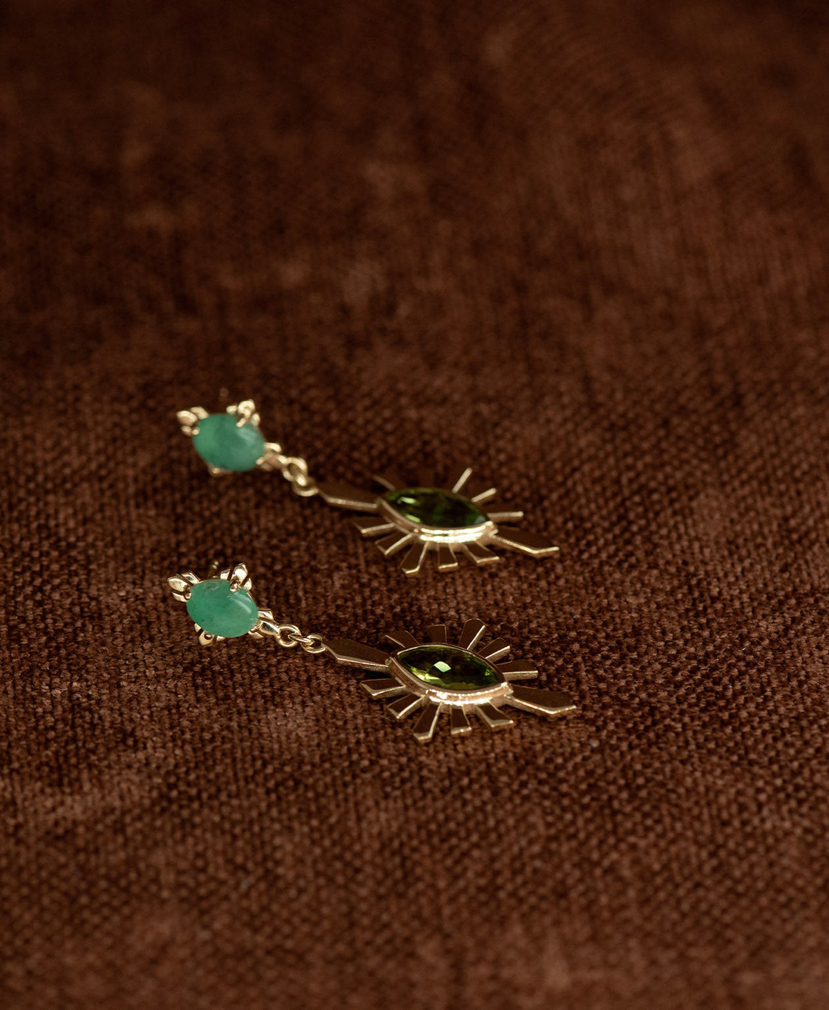 A pair of gold Sisterhood Earrings with a cabochon cut Emerald, set within a linear border, paired with a marquise cut Peridot nestled with a sunburst frame, displayed against a textured brown fabric background.