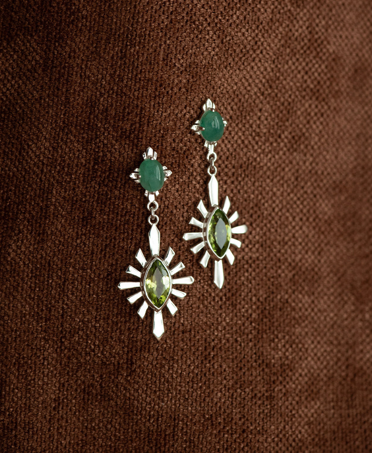 A pair of silver Sisterhood Earrings with a cabochon cut Emerald, set within a linear border, paired with a marquise cut Peridot nestled with a sunburst frame, hanging against a textured brown fabric background.