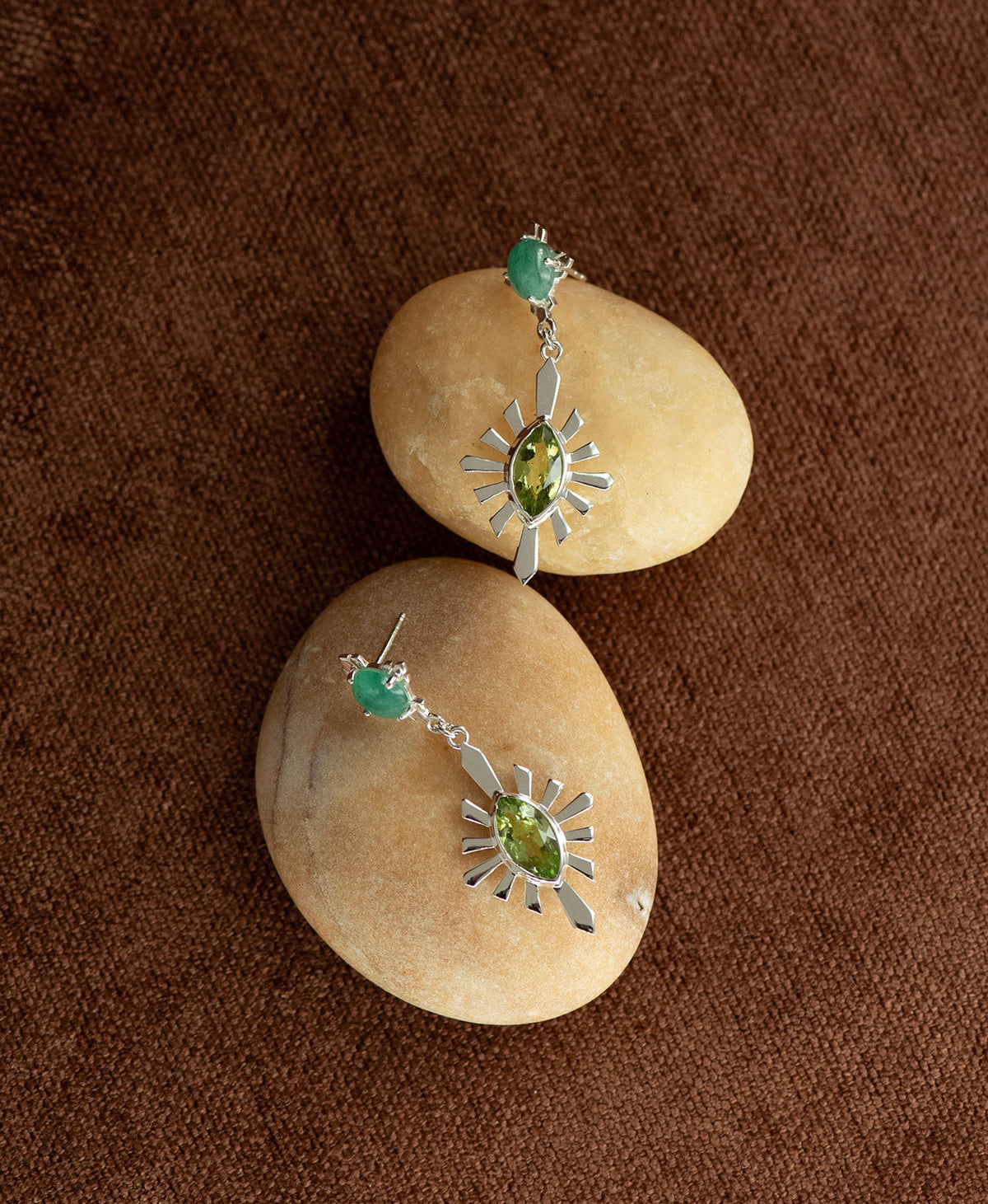 A pair of silver Sisterhood Earrings with a cabochon cut Emerald, set within a linear border, paired with a marquise cut Peridot nestled with a sunburst frame, displayed on a smooth beige stone against a textured brown fabric background.
