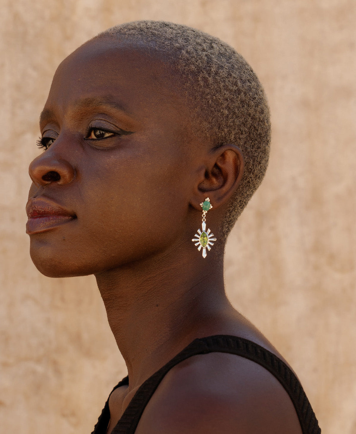 profile of a person wearing the silver Sisterhood Earring, featuring a cabochon cut emerald set within a linear border, paired with a marquise cut peridot with a sunburst frame.