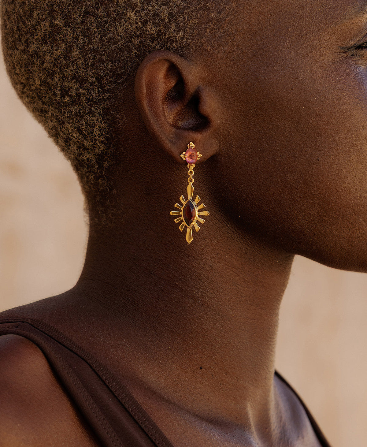 A close-up side profile of a person wearing the Sisterhood Earring, featuring a cabochon cut pink Tourmaline is set within a linear border, paired with a marquise cut Garnet with a sunburst frame.