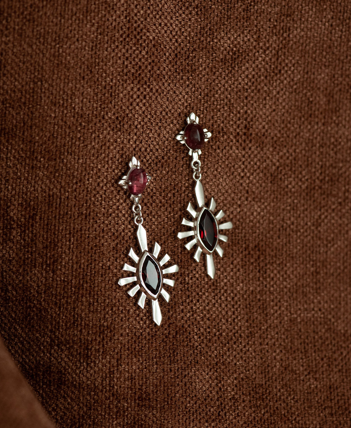 A pair of silver Sisterhood Earrings with a cabochon cut pink Tourmaline, set within a linear border, paired with a marquise cut Garnet nestled with a sunburst frame, hanging against a textured brown fabric background.