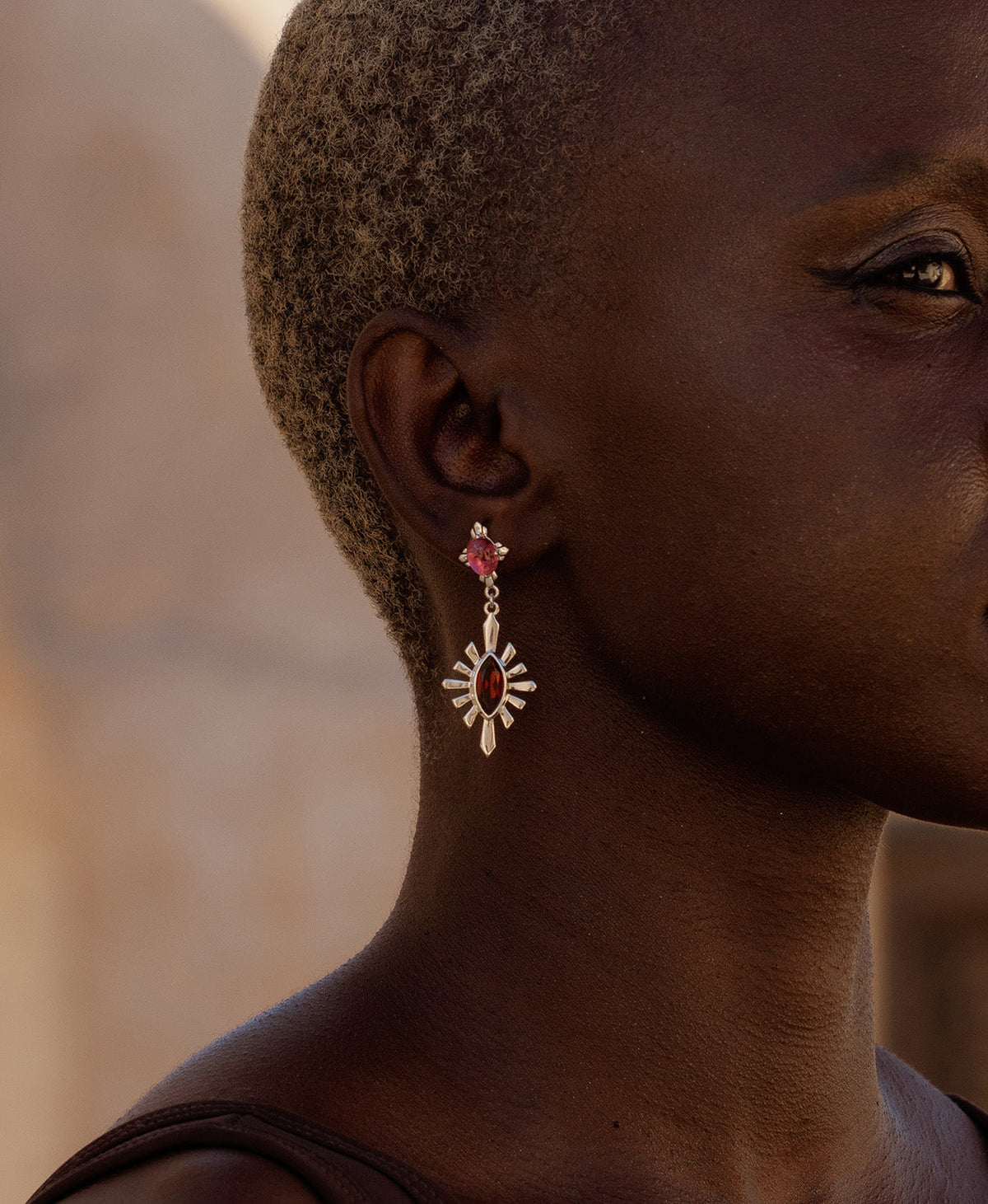 A close-up side profile of a person wearing the silver Sisterhood Earring, featuring a cabochon cut pink Tourmaline is set within a linear border, paired with a marquise cut Garnet with a sunburst frame.