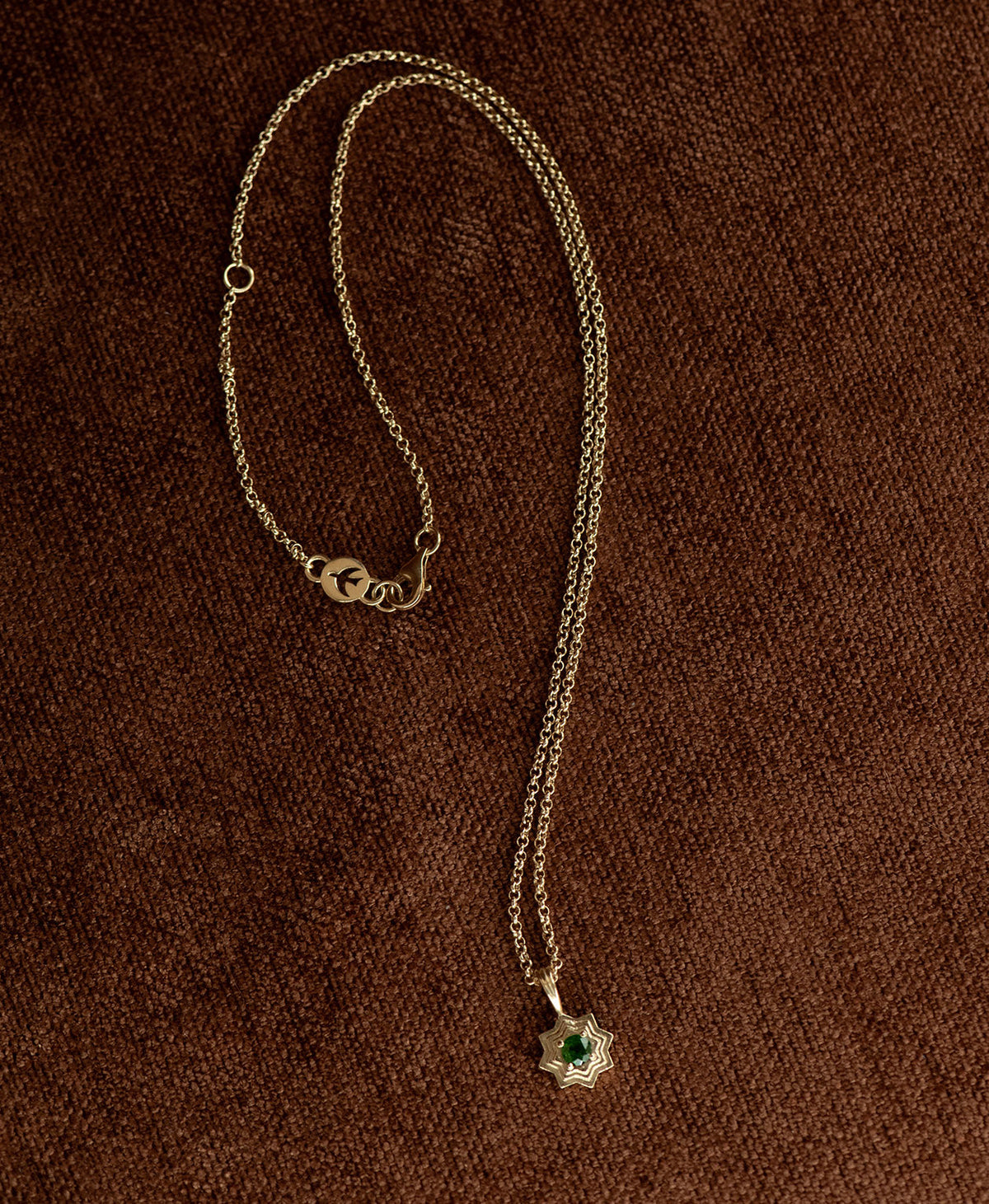 The Sitara Necklace featuring a gold star like pendant with concentric lines that radiate outwards from a Chrome Diopside resting at its centre, displayed on a textured brown fabric background.