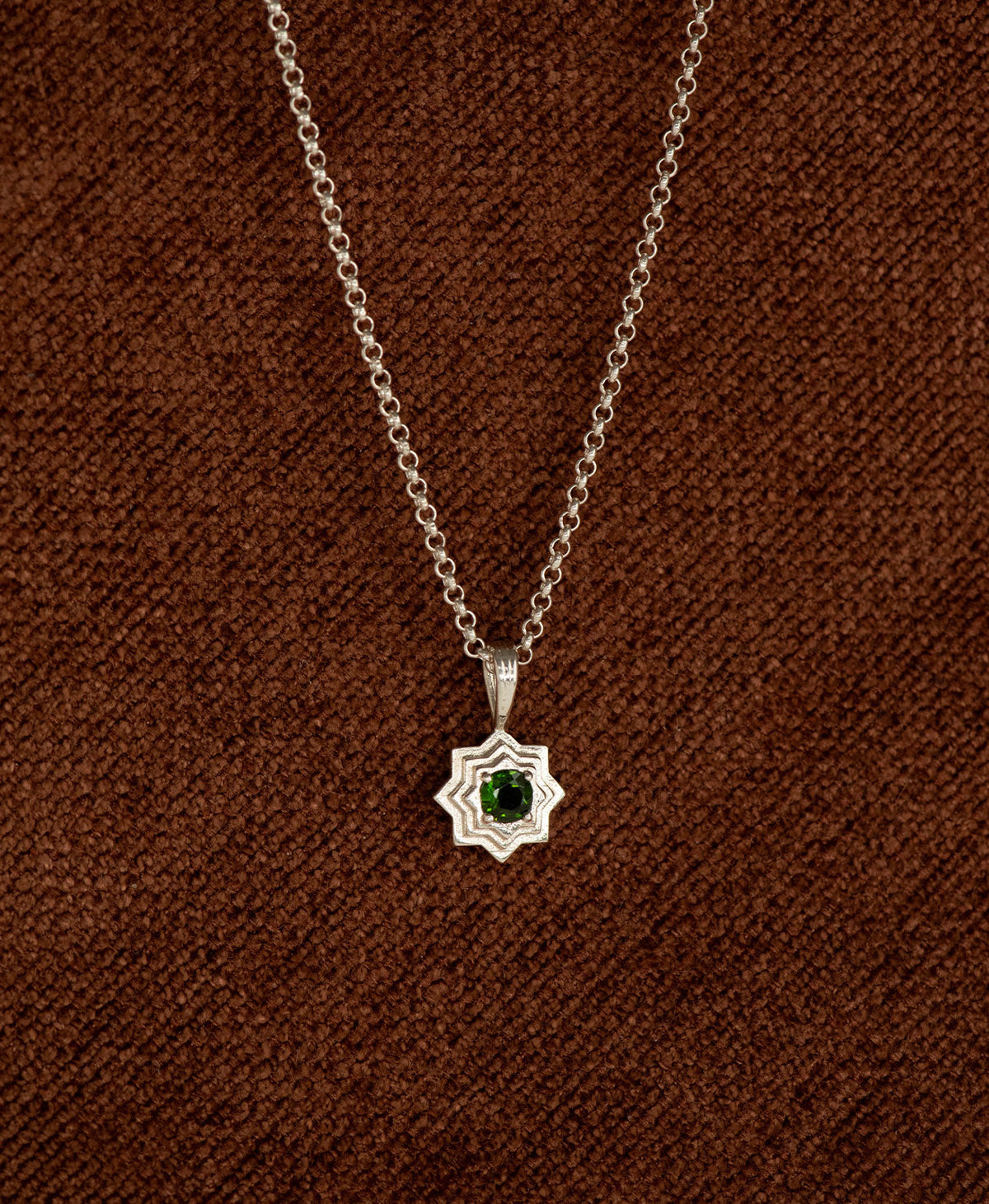 The Sitara Necklace featuring a silver star like pendant with concentric lines radiating outwards from a Chrome Diopside resting at its centre, displayed on a textured brown fabric background.
