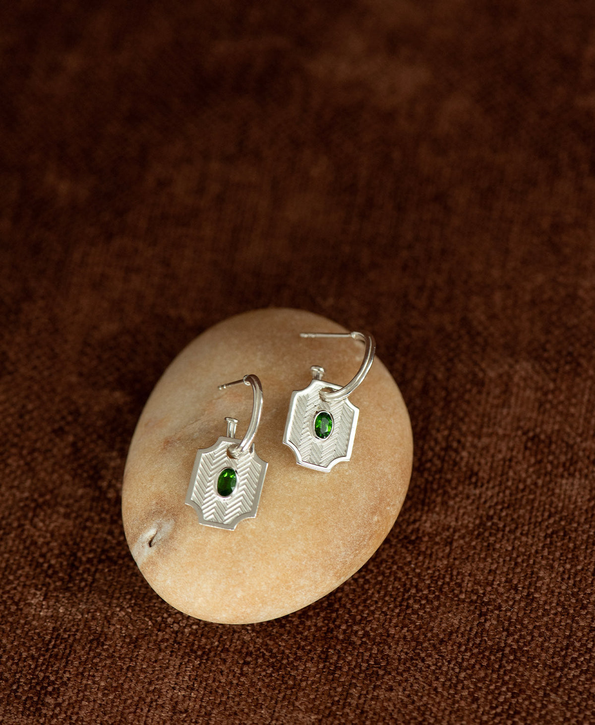 The Soul Sister Hoops, featuring a silver geometric pendant threaded onto a hoop, displayed on a smooth beige stone against a textured brown fabric background.