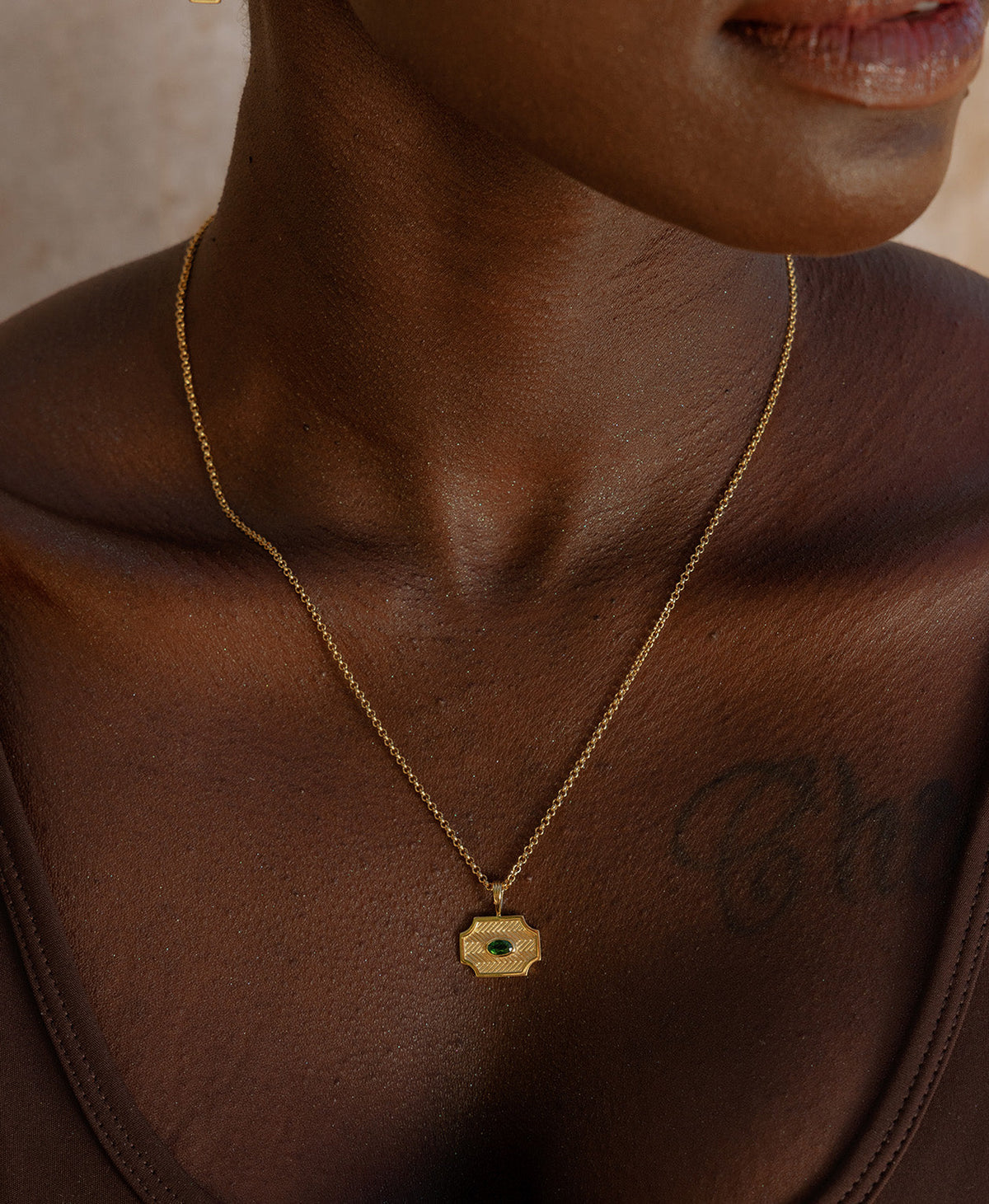 A close-up of a neck wearing the Soul Sister Necklace, a gold textured pendant with a central green stone, suspended from a fine belcher chain.