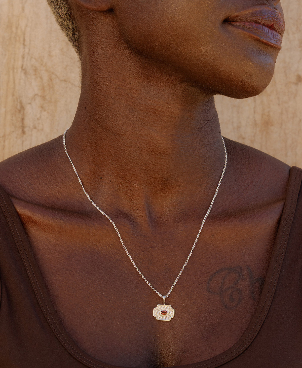 A close-up of a neck wearing the Soul Sister Necklace, a silver textured pendant with a central pink stone, suspended from a fine belcher chain.
