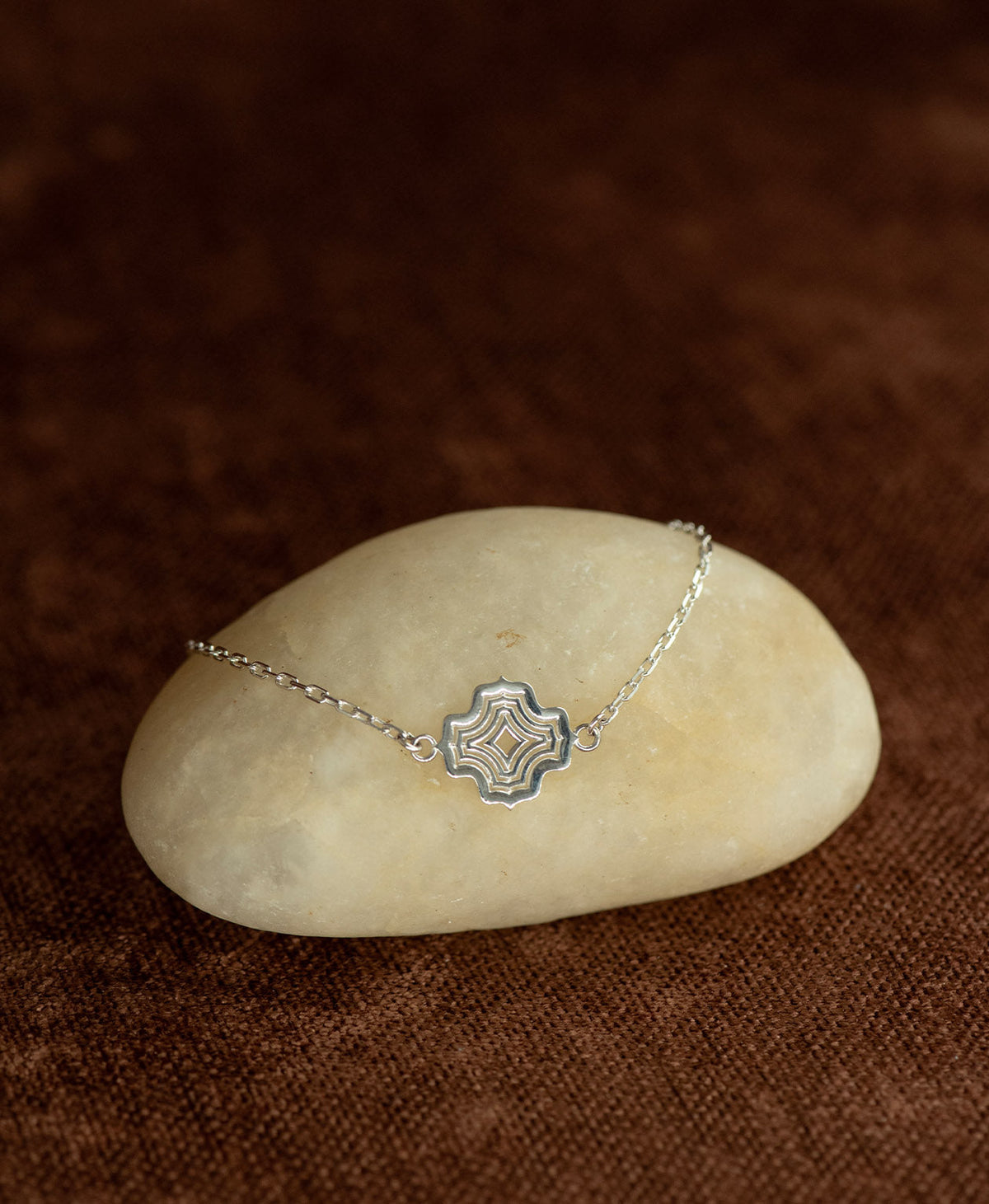 The Alma Bracelet featuring a silver pendant with a concentric wave design, layered over a cream stone on a textured brown fabric background.