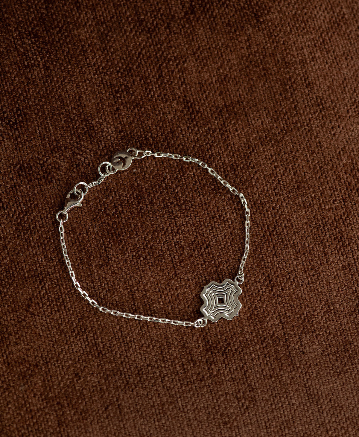The Alma Bracelet featuring a silver pendant with a concentric wave design, on a textured brown fabric background.