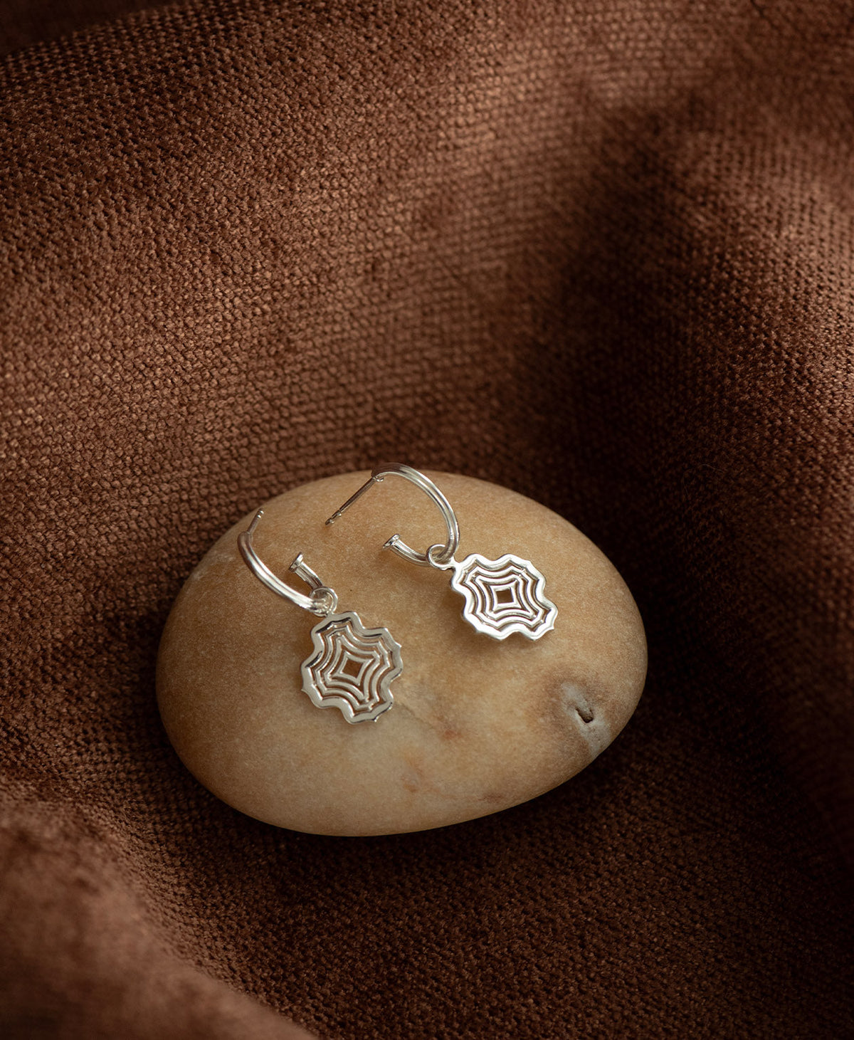 The Alma Hoops, featuring a silver pendant with a concentric wave design suspended from a hoop, displayed on a smooth beige stone against a textured brown fabric background.