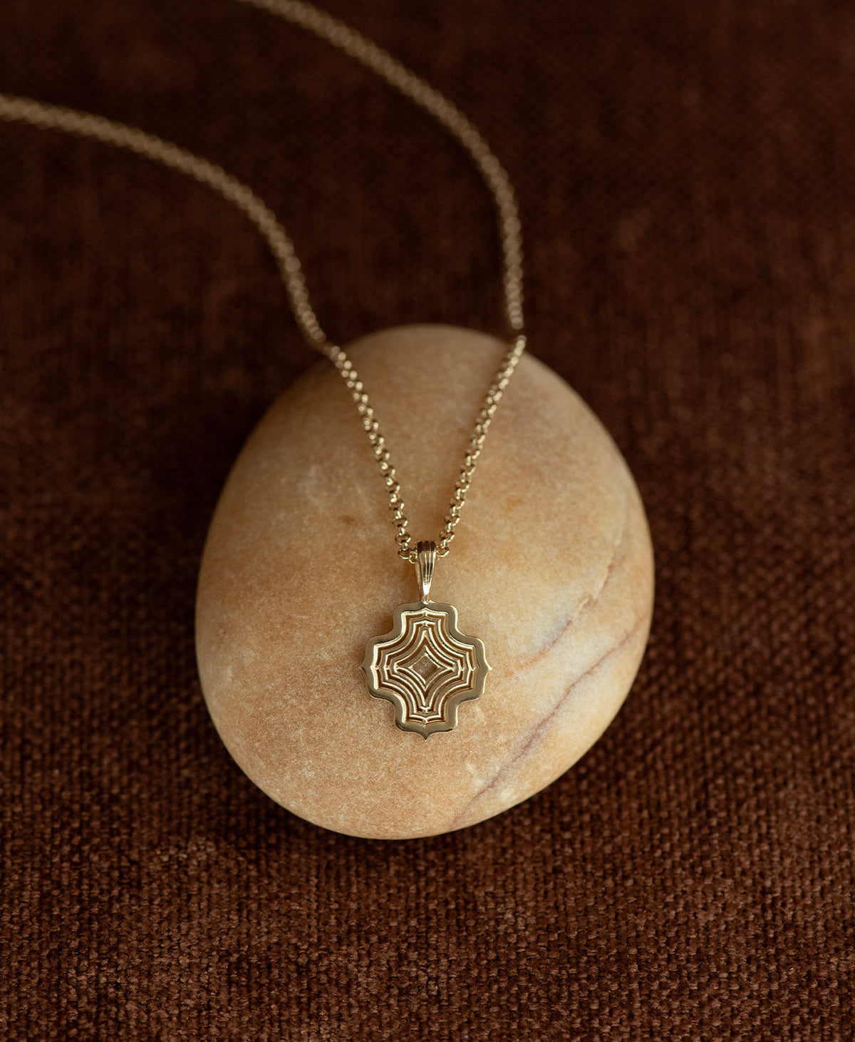 The Alma Necklace featuring a gold pendant with a concentric wave design, layered over a cream stone on a textured brown fabric background.