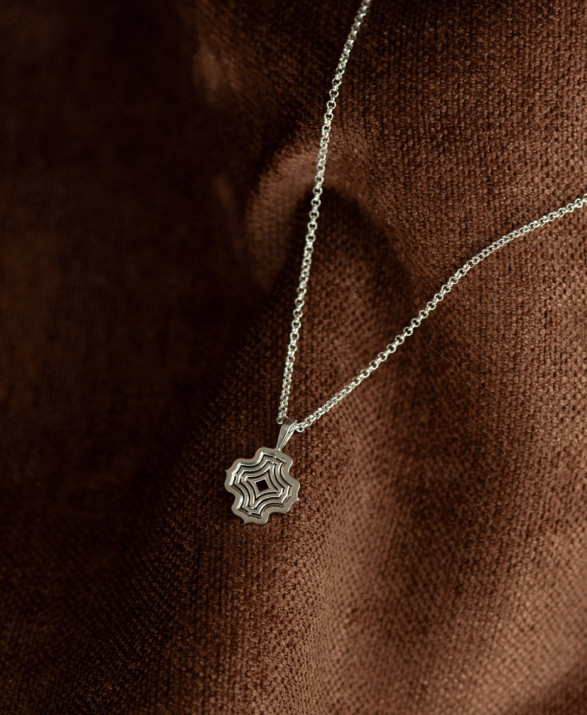 The Alma Necklace featuring a silver pendant with a concentric wave design, displayed on a textured brown fabric background.