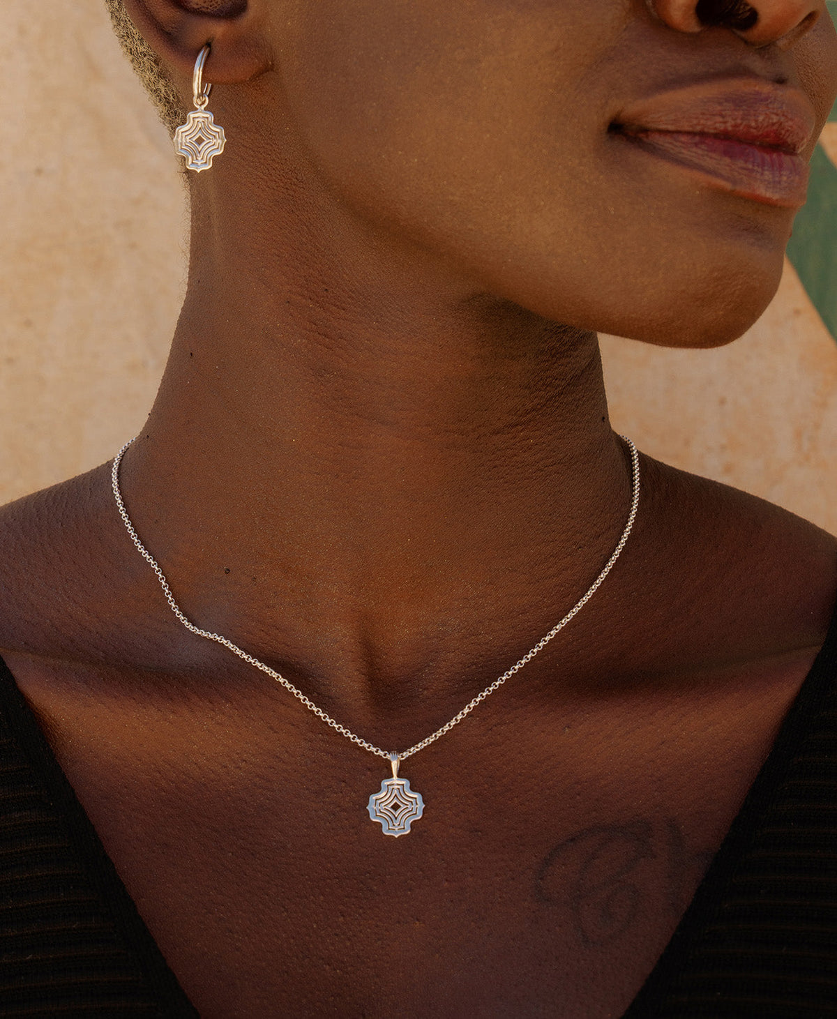 A close-up of a neck wearing the Nora Necklace, a silver pendant with a concentric wave design, suspended from a fine belcher chain, paired with the matching silver earrings. 