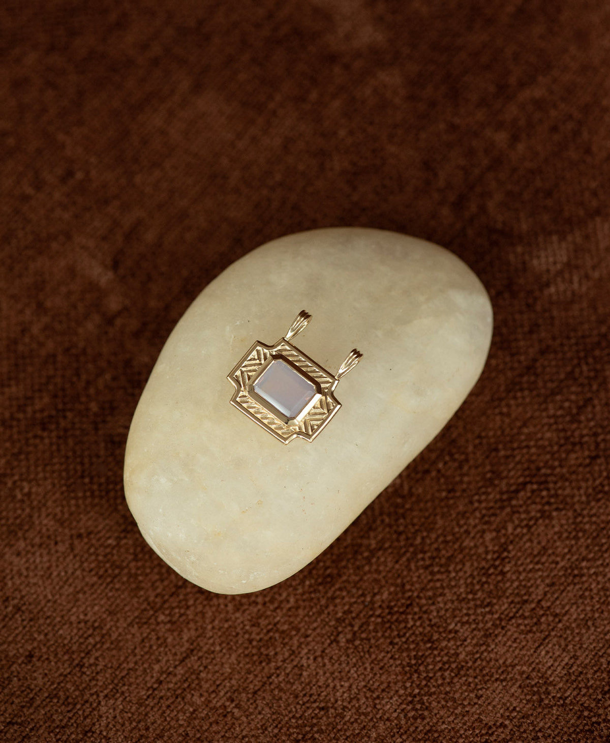 A gold pendant with a rectangular lavender quartz stone, on a smooth white stone on a textured brown background.