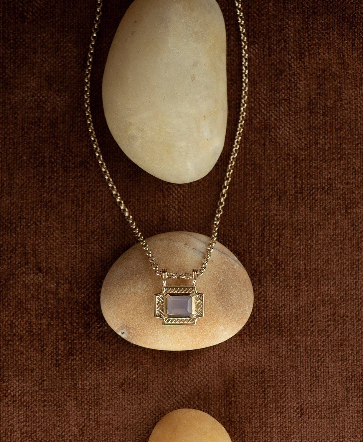 A gold pendant with a rectangular lavender quartz stone, handing on a belcher chain handing on a cream stone against a textured brown background.
