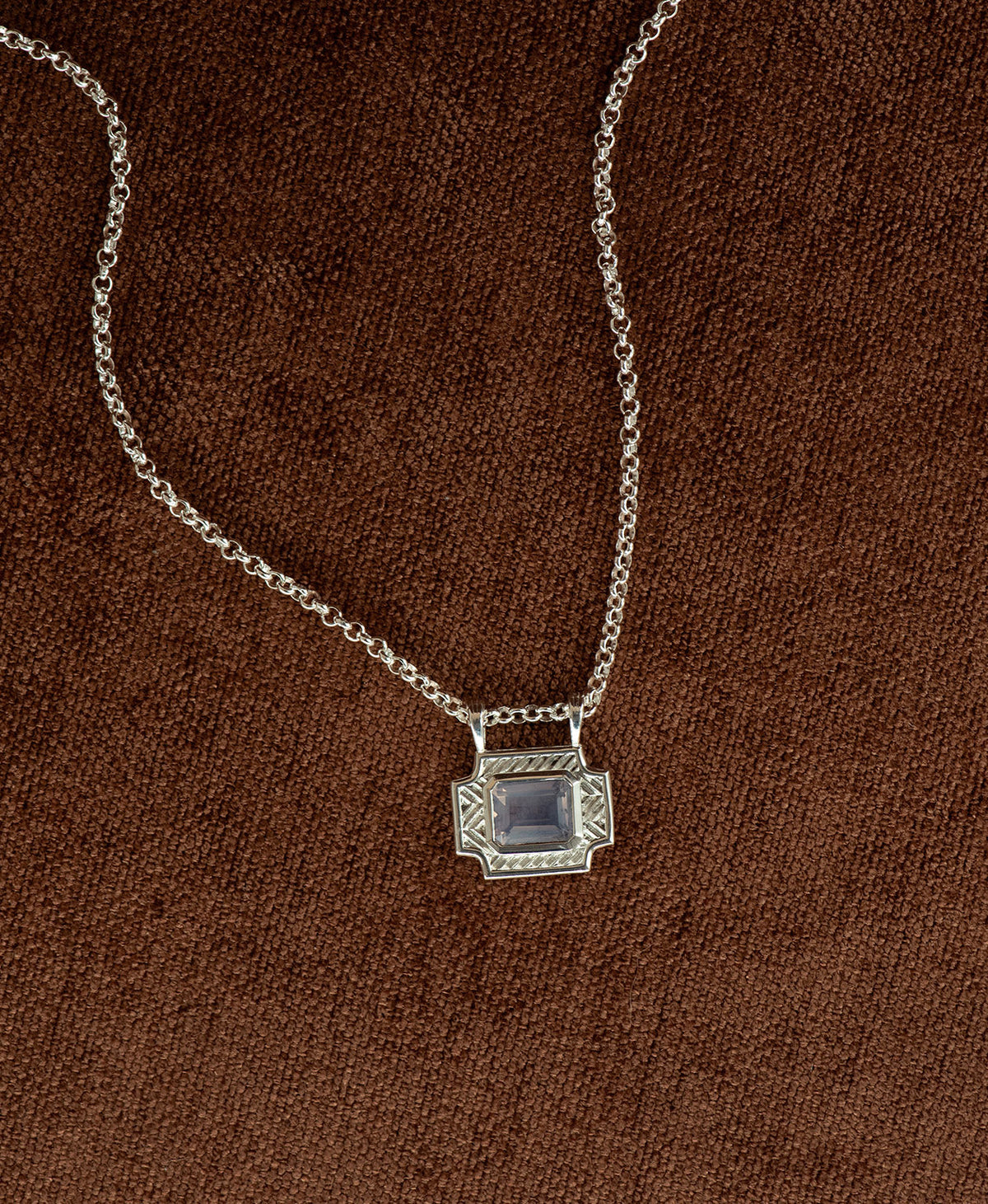 A silver pendant with a rectangular lavender quartz stone, hanging on a belcher chain against a textured brown background.