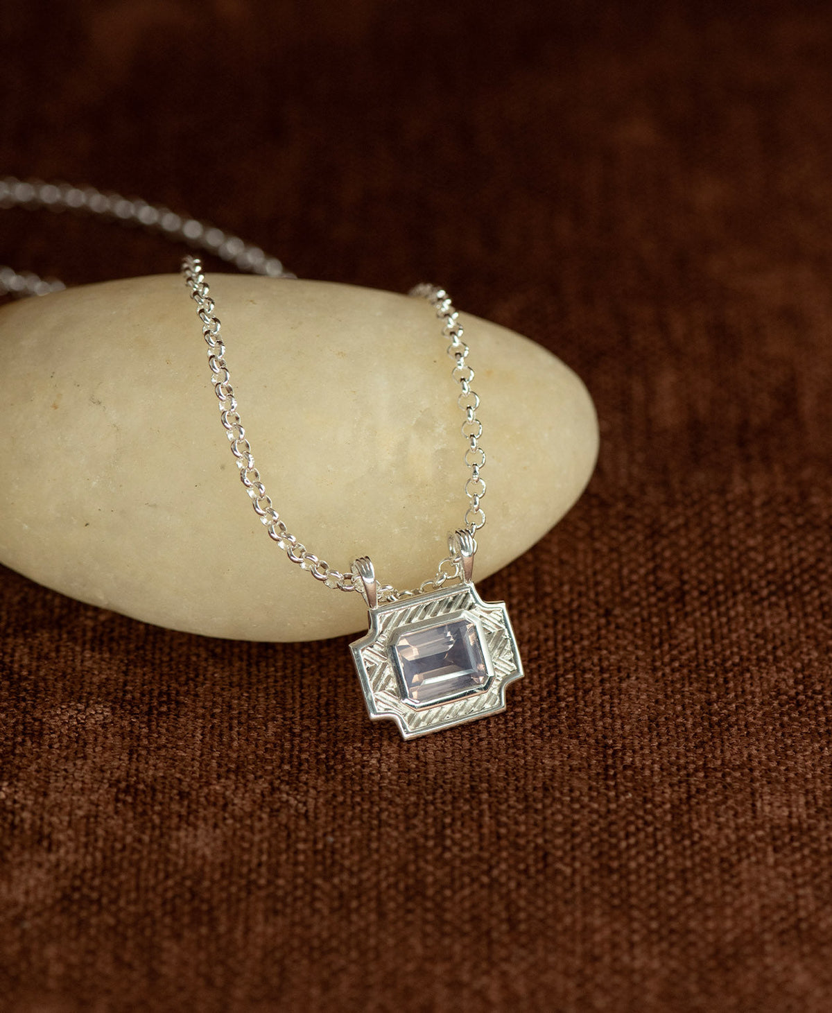 A silver pendant with a rectangular lavender quartz stone, resting on a cream stone hanging on a belcher chain against a textured brown background.