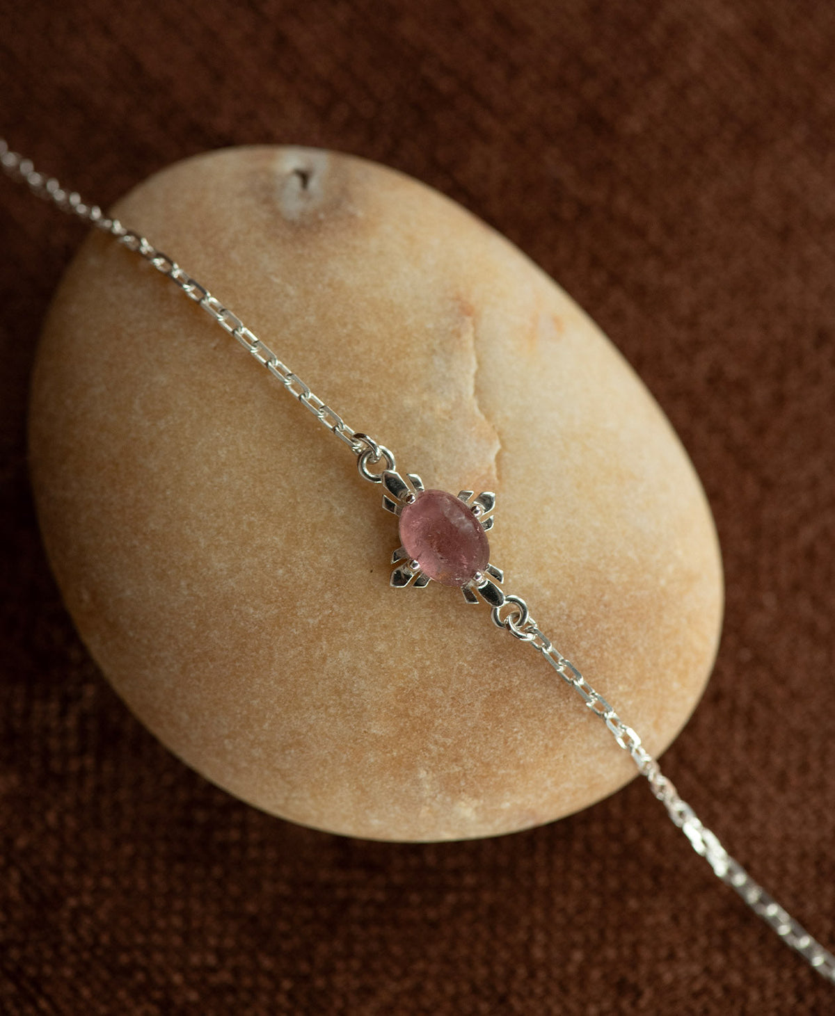A close up of the silver Farah Bracelet featuring a cabochon cut pink tourmaline, layered over a cream stone on a textured brown fabric background.