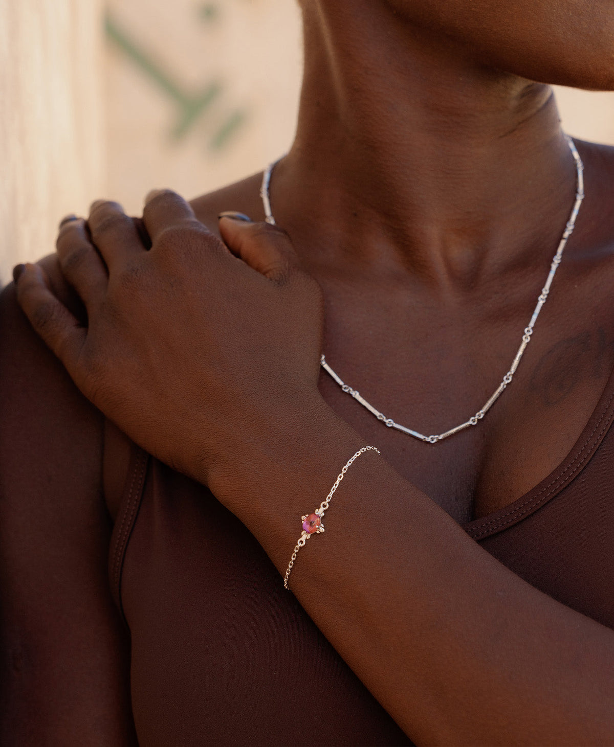 A close-up of a person wearing the Farah Bracelet, a silver pendant with a cabochon cut pink tourmaline, set on a fine cable chain, paired with a mid length silver chain necklace. 