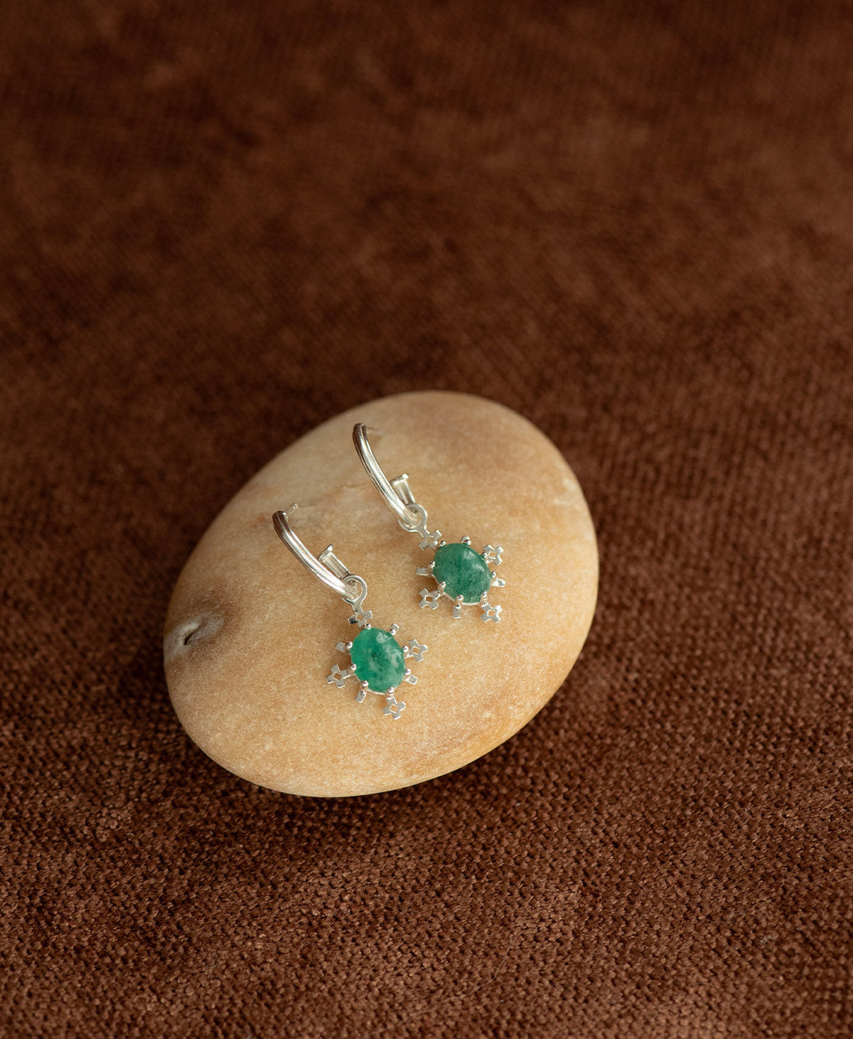 A pair of Farah silver drop earrings with emerald gemstones, displayed on a smooth cream coloured stone against a dark brown textured background.