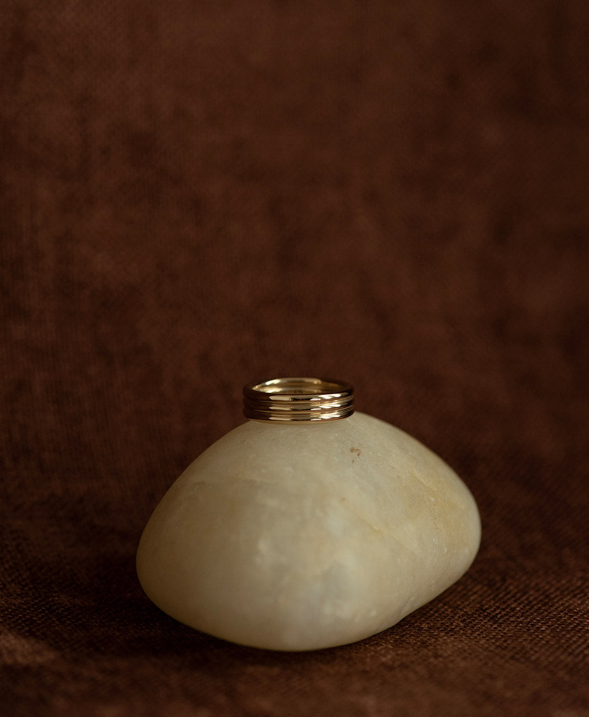 The gold Irma Stacker Ring, formed from three connected bands, layered closely, rests on a cream stone on a textured brown background.