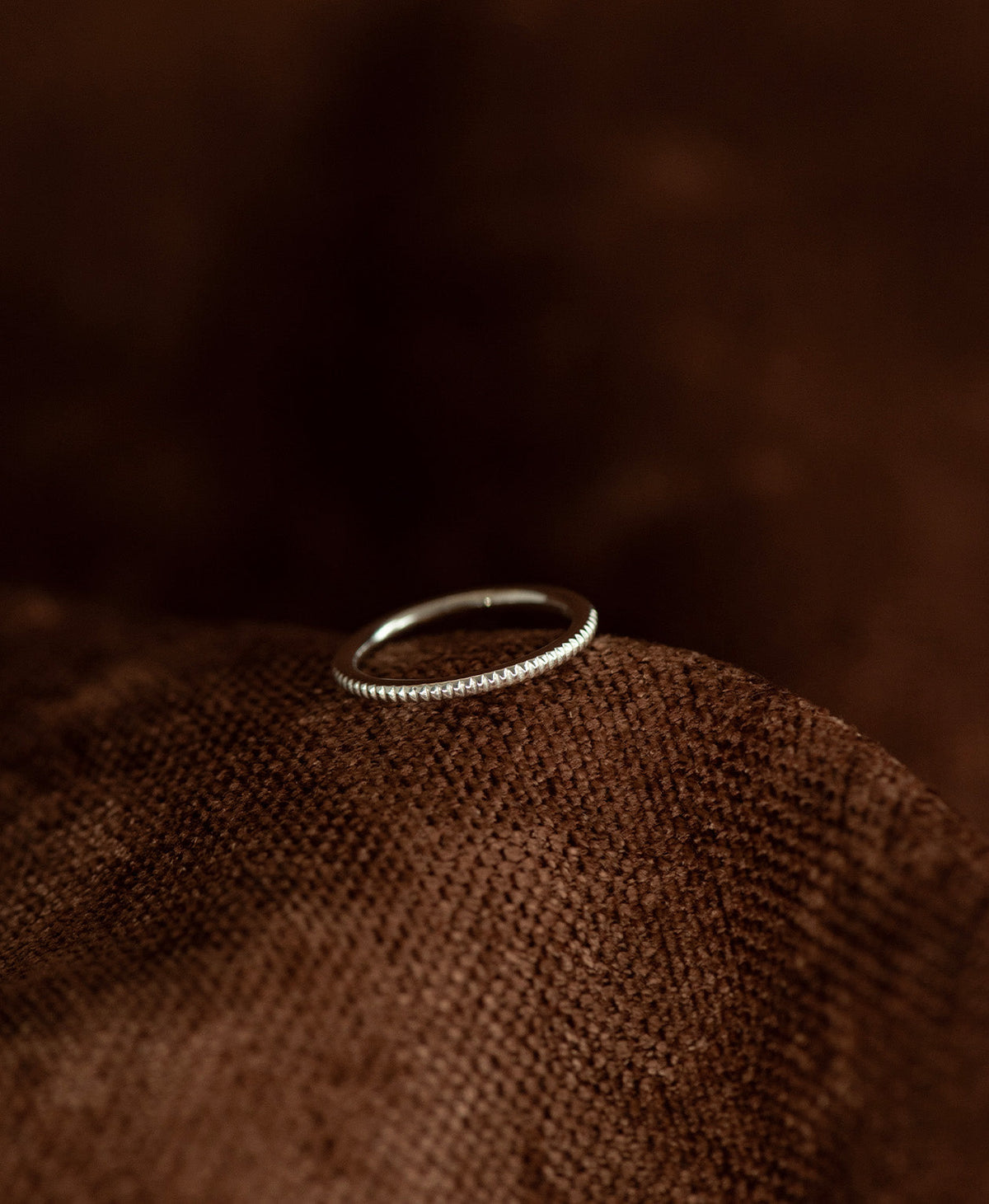 The silver Kenza Stacking Ring with fine engraved lines encircling the band, rests on a textured brown background.
