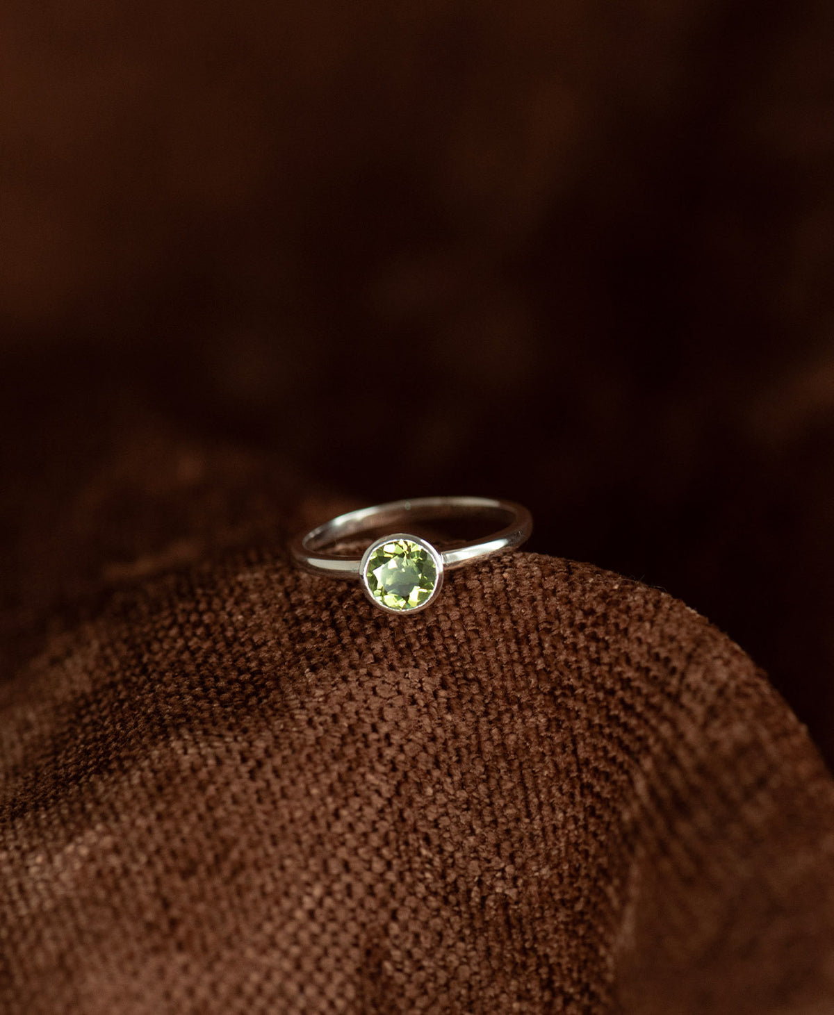 A close up of the silver Mon Petit Stacker Ring with a bezel set, round cut Peridot resting on a textured brown background.