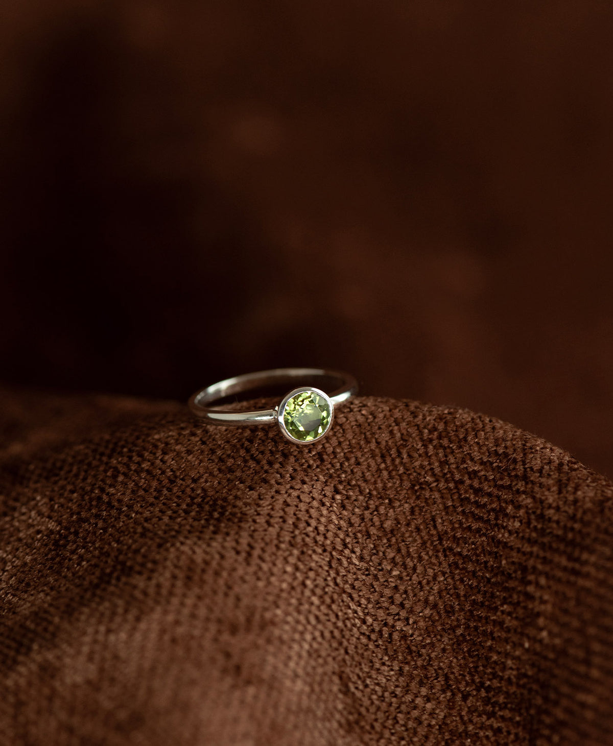 The silver Mon Petit Stacker Ring with a bezel set, round cut Peridot rests on a textured brown background.