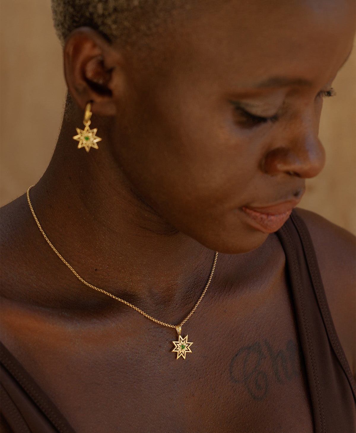A side profile of a person wearing the Nora Necklace, a gold eight pointed star pendant with a central green stone, paired with the matching gold earrings.  