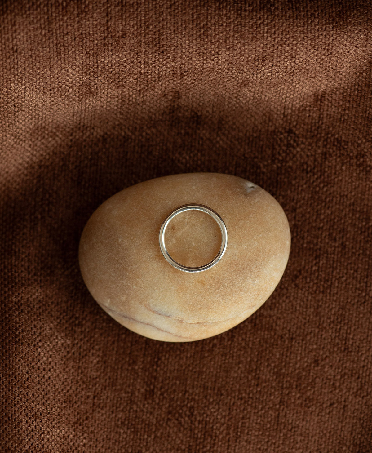 The smooth, curved Selma stacking ring in 925 sterling silver, lays on a cream stone on a textured brown background. 