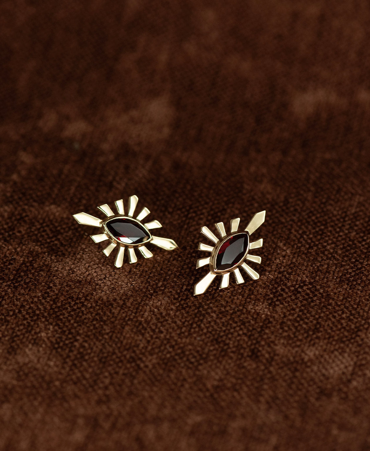 The Sister Studs, featuring a central marquise cut Garnet nestled within a gold sunburst frame, displayed against a textured brown fabric background.